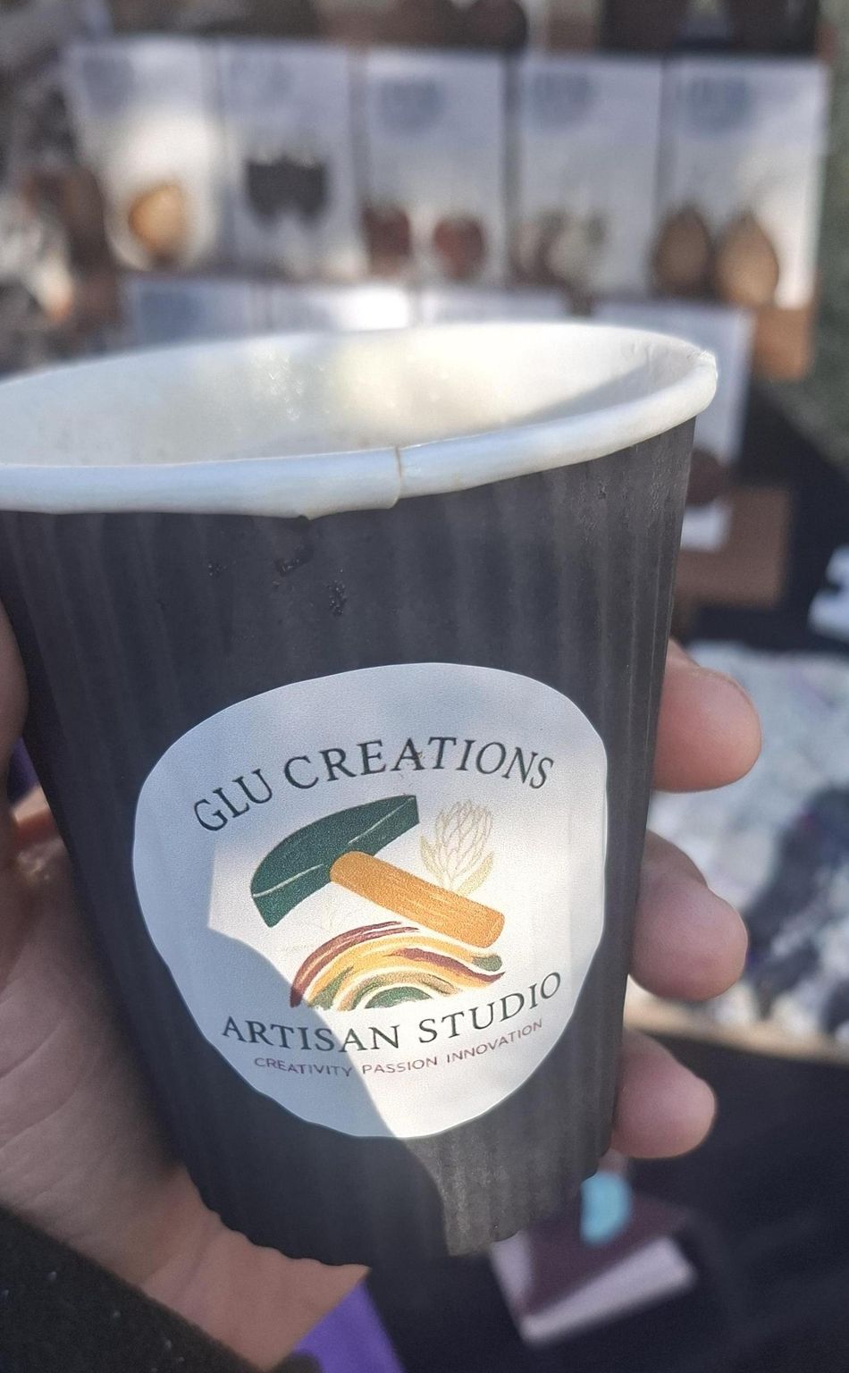 A coffee cup with the GLU Creations Artisan Studio logo, held at a market stall with leather earrings displayed behind it